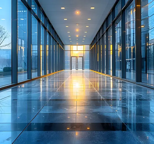 Modern glass hallway with reflective tiled floor and ceiling lights, sun setting at the glass door end.