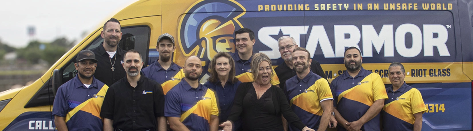 Group of twelve smiling people standing in front of a vehicle branded with STARMOR logo and text about safety and riot glass.