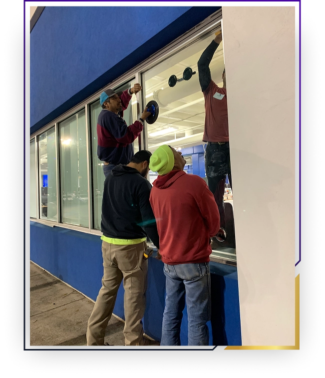 Four men installing or cleaning a large glass window on a blue building at night.