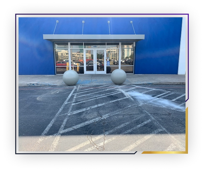 Entrance to a building with blue walls, glass double doors, and two large spherical concrete bollards in front on a parking lot with marked lines.