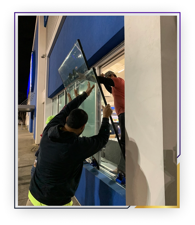 Two men installing or removing a large glass window pane at night on a blue building.