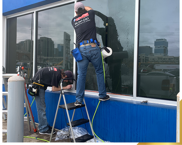 Two workers cleaning large windows on a building exterior using squeegees and water with city buildings reflected in the glass.