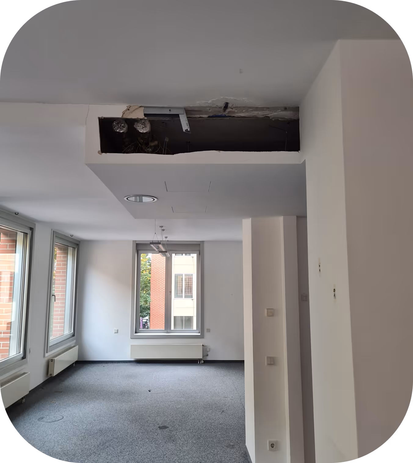Leerer Raum mit grauem Teppichboden, weißen Wänden, mehreren Fenstern und öffentlicher Decke mit sichtbarem Rohrleitungsabschnitt.