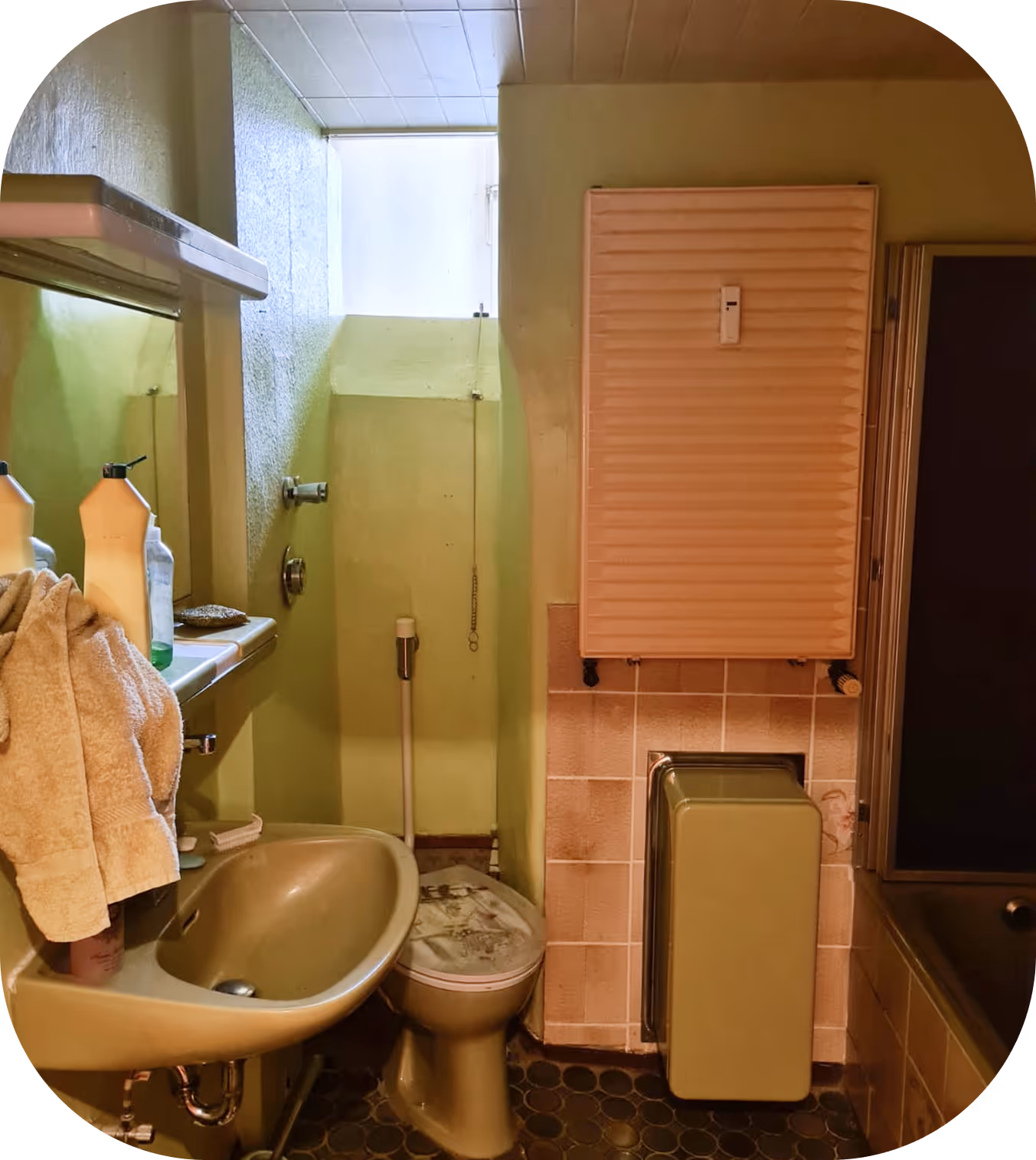 Kleines Retro-Badezimmer mit grünem Waschbecken, Toilette, Handtuch an Waschbecken, Heizkörper und kleinem Fenster.