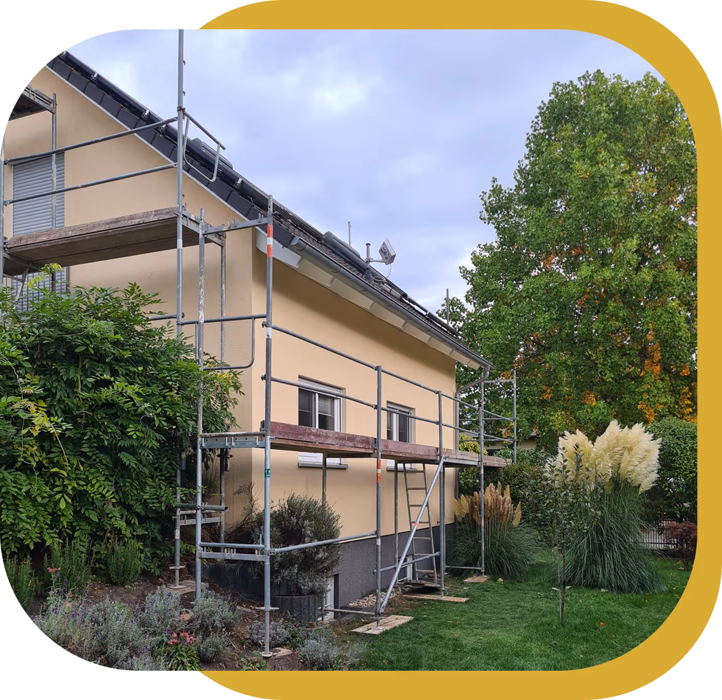 Haus mit gelben Wänden und Gerüst zur Fassadenarbeit, umgeben von grünem Garten mit Bäumen und Ziergras.