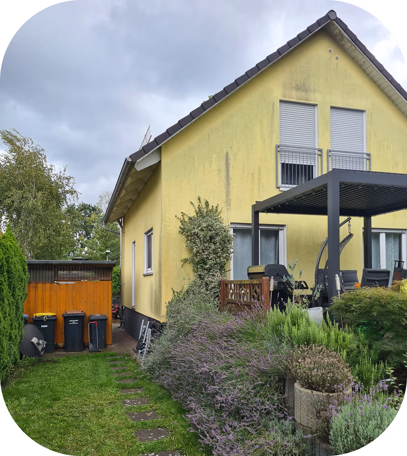 Gelbes Haus mit geschlossener Terrasse, Garten mit lila blühenden Sträuchern und Mülltonnen neben Holzschuppen.