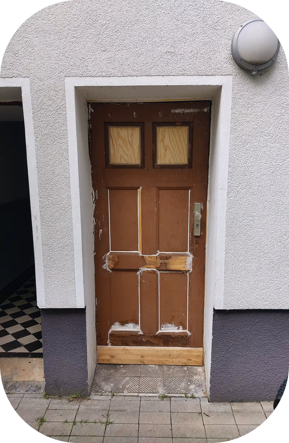 Braune Holztür mit beschädigten Stellen und Reparaturen an einer weißen Hausfassade neben einem Flur mit schwarz-weiß kariertem Boden.