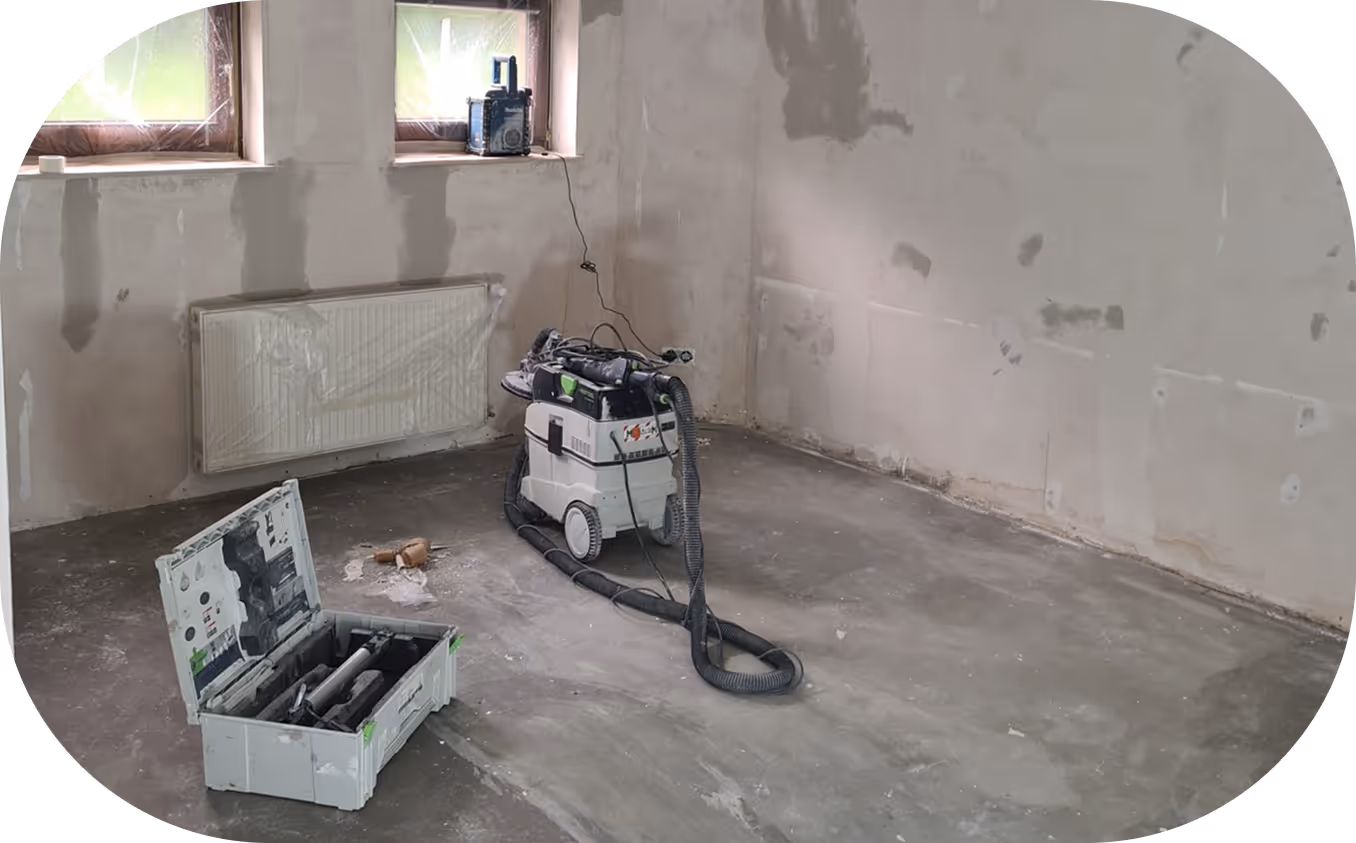 Unfertiger Raum mit verputzten Wänden, zwei Fenstern, radiator und geöffnetem Werkzeugkoffer sowie einer Staubsaugeranlage auf dem Boden.