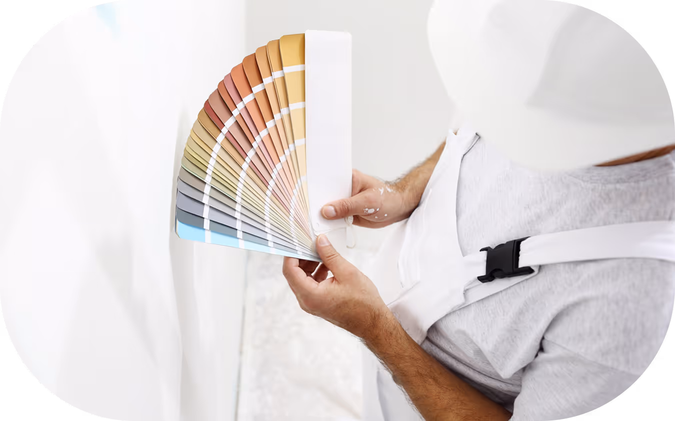 Mann in weißem Maleranzug und Helm hält einen Farbfächer mit verschiedenen Wandfarben vor eine weiße Wand.