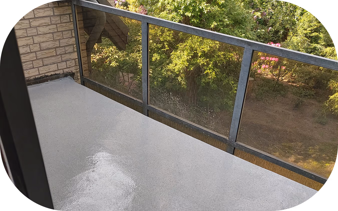 Balkon mit feuchtem, glänzendem Bodenbelag und Glasgeländer mit Blick auf grüne Bäume und Blumen.