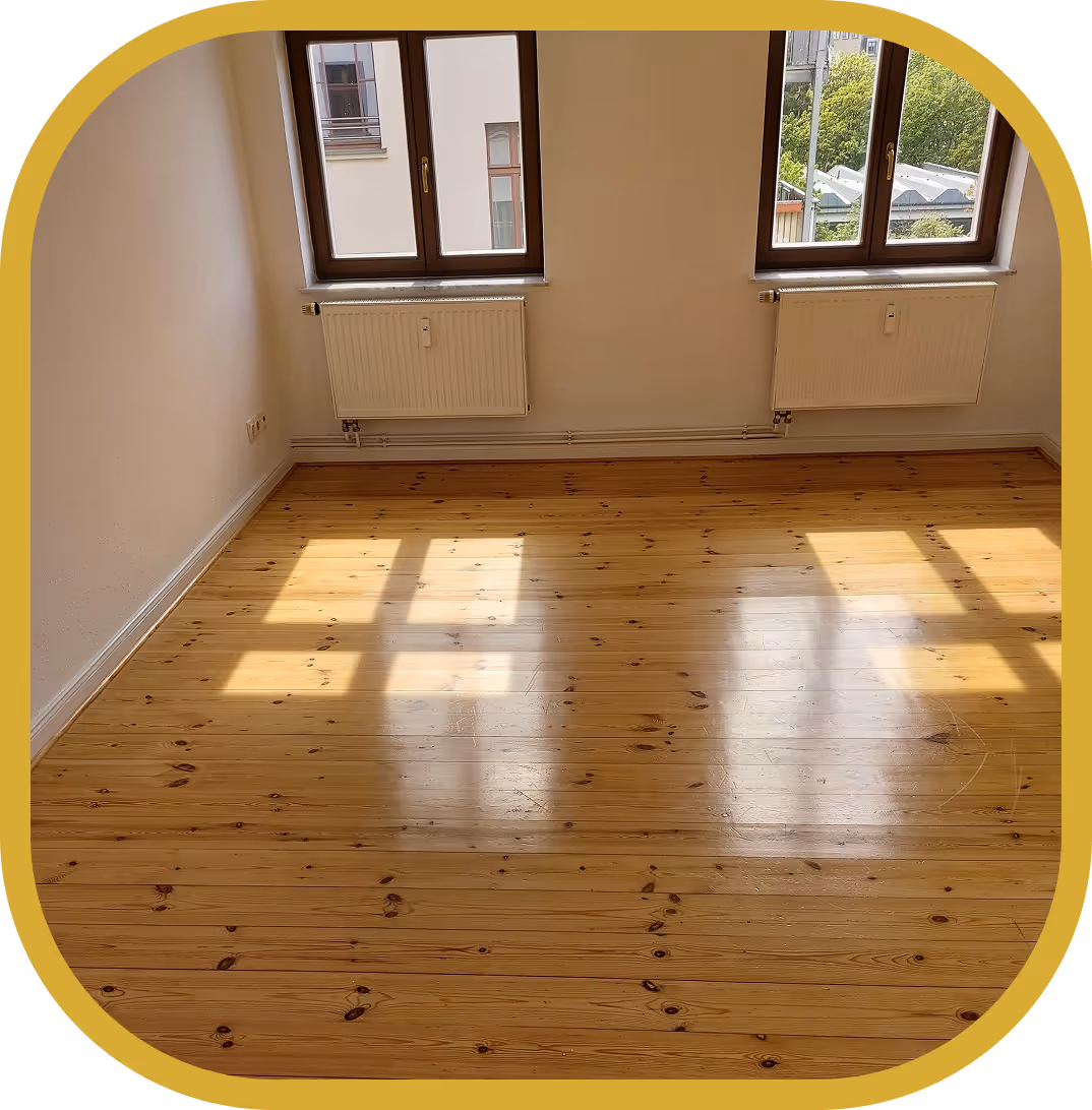 Leerer Raum mit zwei Fenstern, Holzboden und weißen Heizkörpern unter den Fenstern.