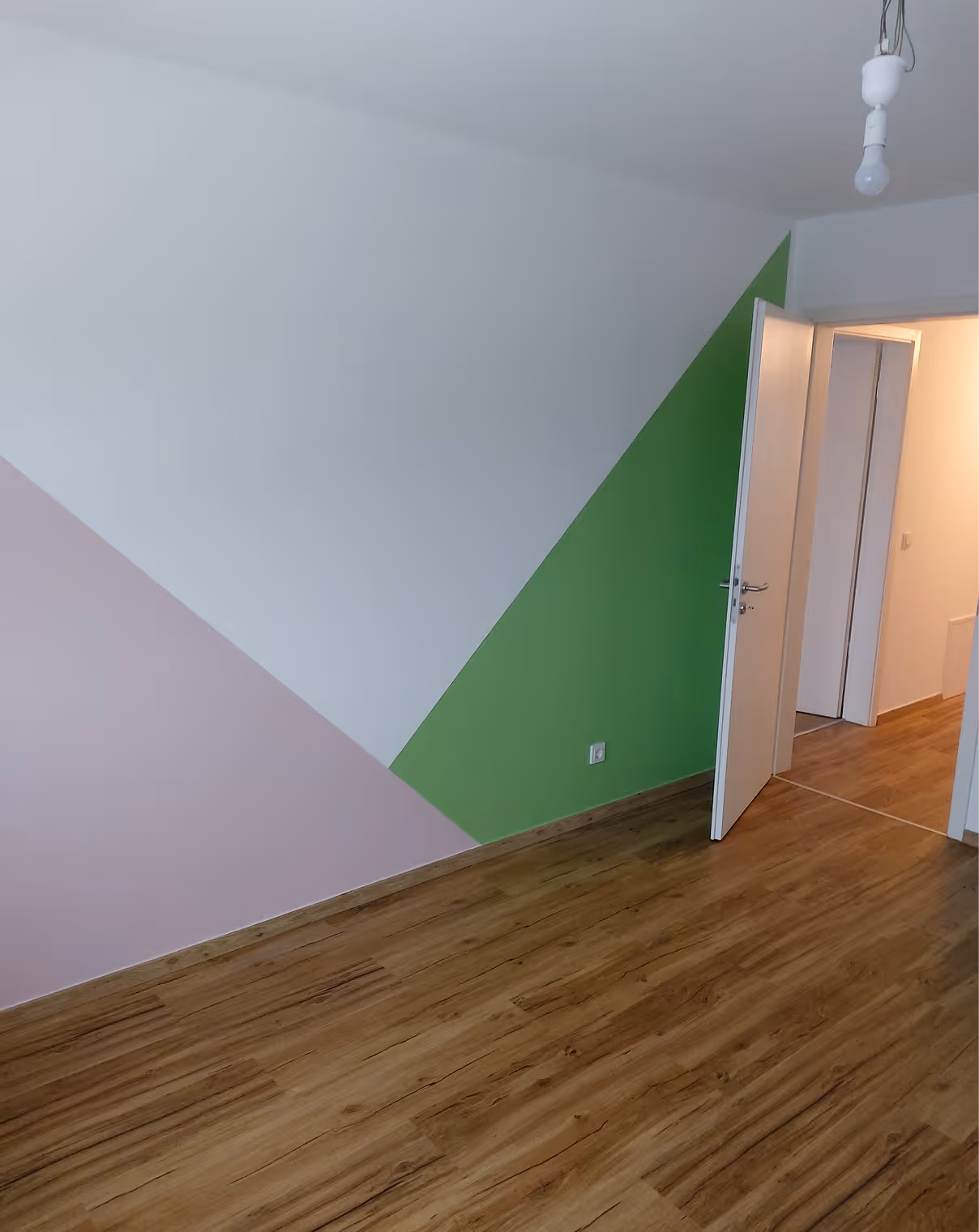 Zimmer mit braunem Holzboden, einer weißen Wand mit grünen und rosa Dreiecken und einer offenen Tür zum Flur.