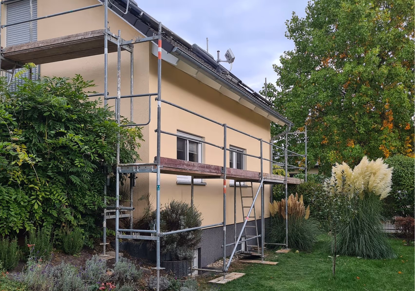 Einfamilienhaus mit gelber Fassade und Metallgerüst für Sanierungsarbeiten, umgeben von grünen Büschen und Rasen.