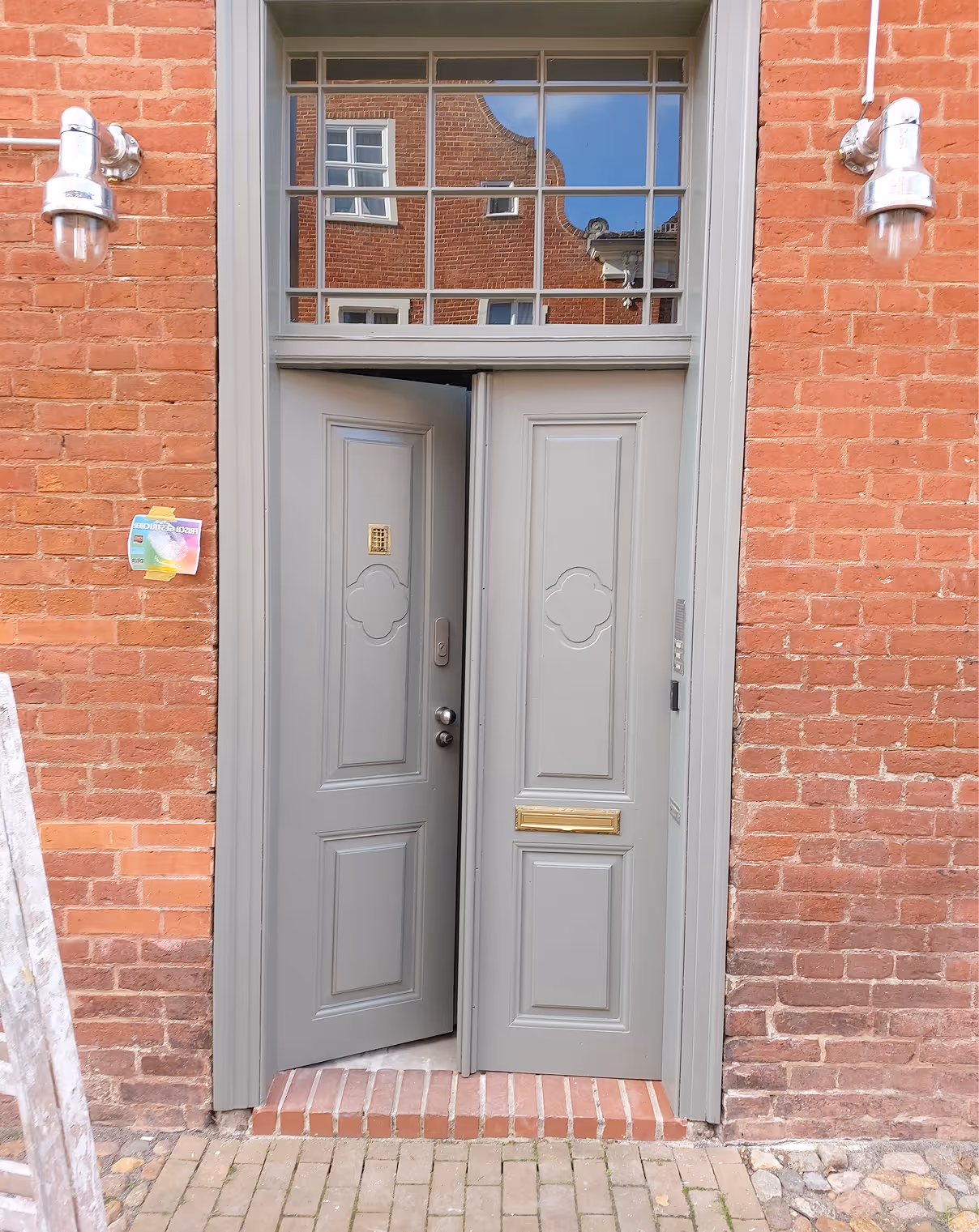 Halb geöffnete graue Doppeltür mit dekorativen Paneelen in einer Ziegelsteinwand mit zwei Wandlampen.