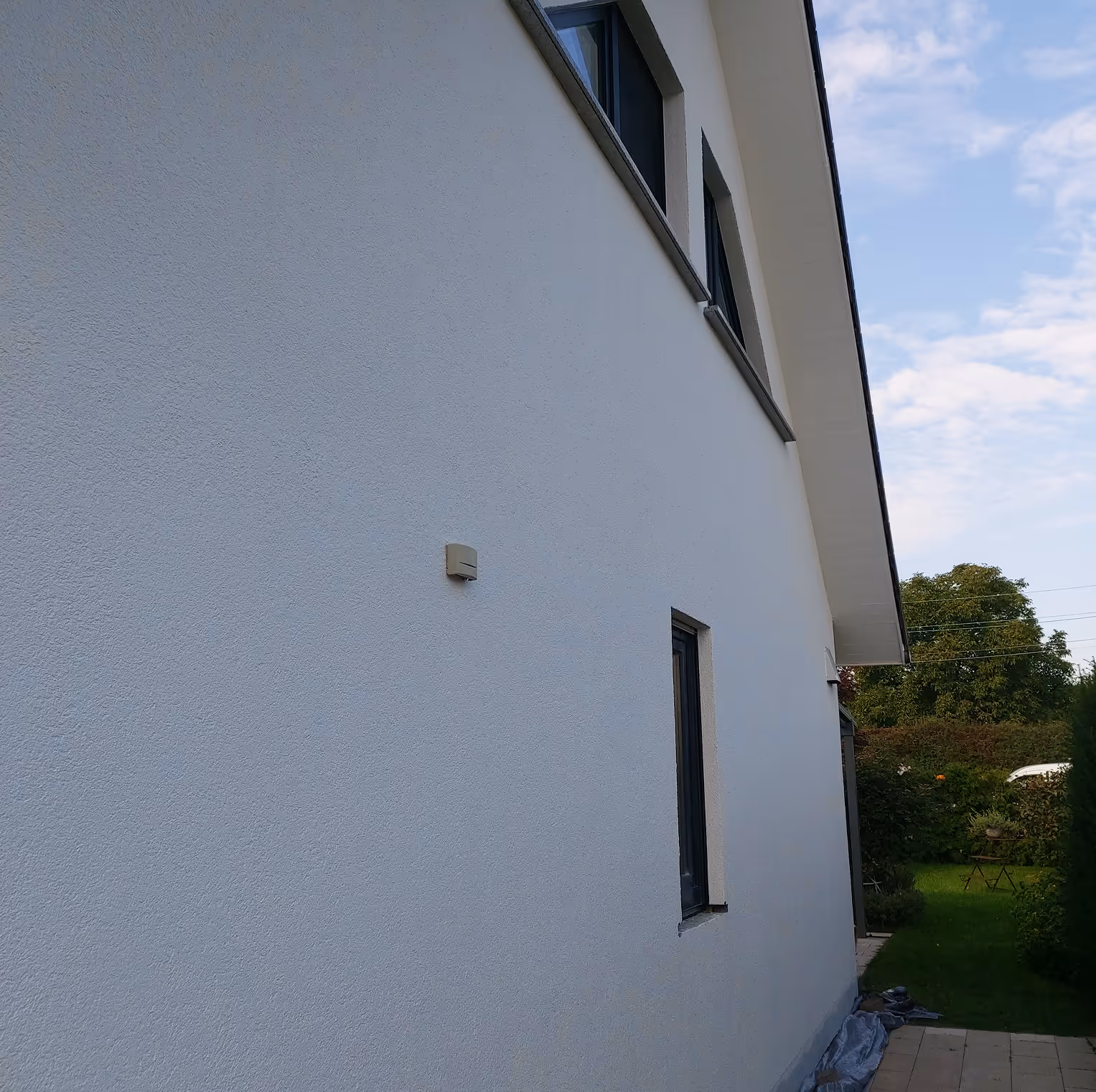 Weiße Hauswand mit zwei kleinen rechteckigen Fenstern, grüner Garten mit Stuhl und Tisch im Hintergrund.