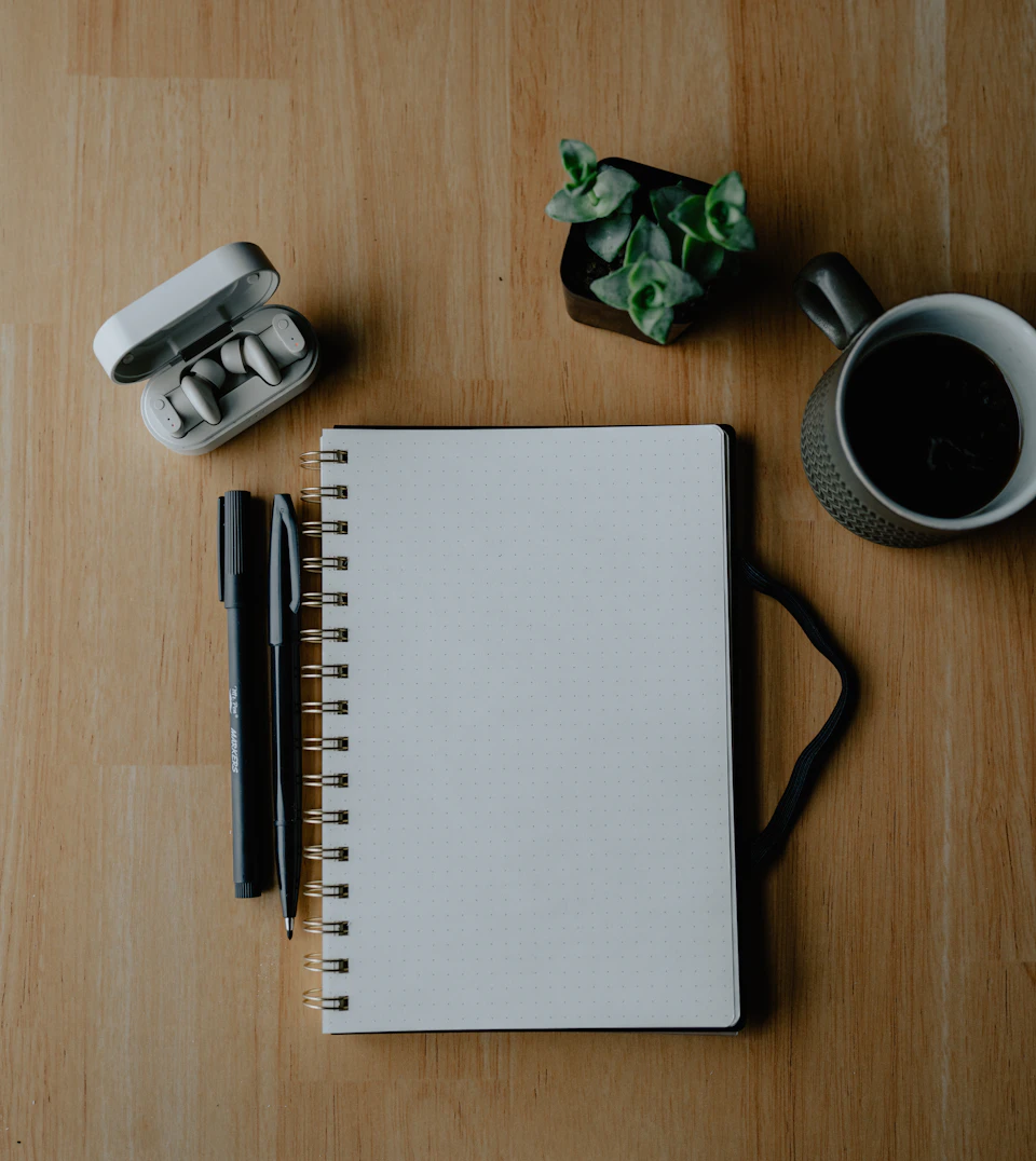 An open notebook, pens and coffee arranged on a wooden desk
