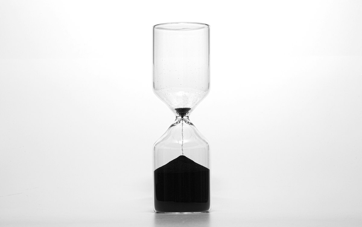 A modern hourglass on a white surface with nearly all of its black sand already in the bottom chamber, a fitting picture of the Data Center calendar public sector customers are planning against. Photo by Wilhelm Gunkel on Unsplash.