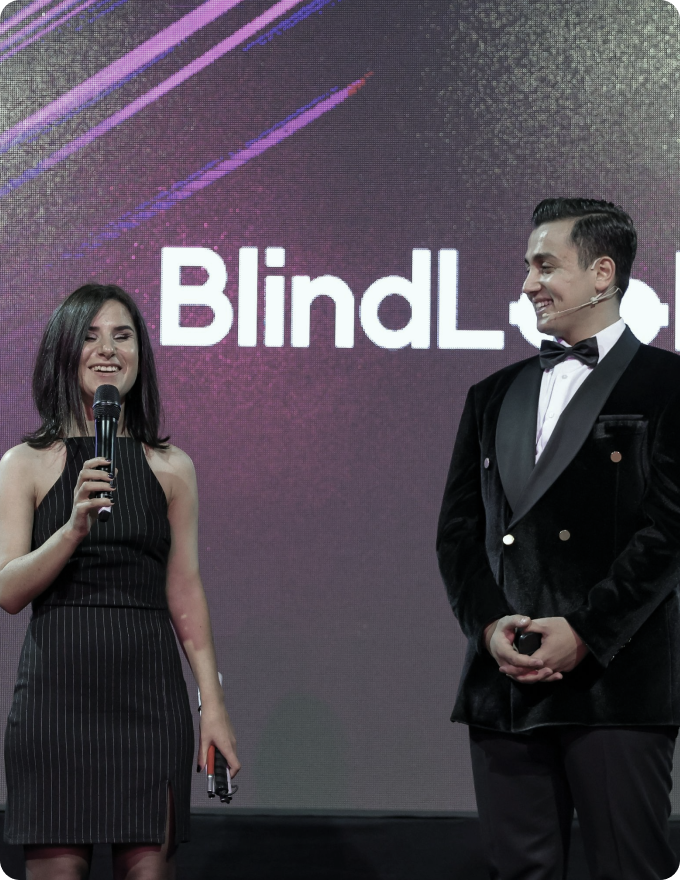 Two presenters speaking on stage in front of a BlindLook event screen.