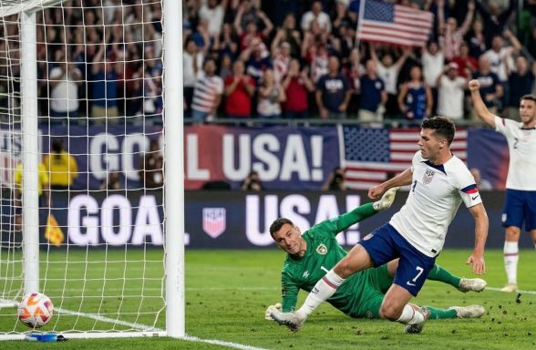 Bir maç sırasında ABD'li futbolcu gol atarken, kaleci kurtarmaya çalışıyor ve arka planda taraftarlar sevinç gösterisinde bulunuyor.