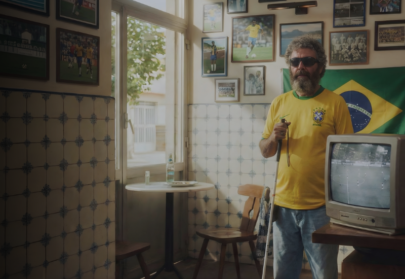 Visually impaired Brazil football fan wearing a Brazil jersey and sunglasses holding a white cane in a bar with a Brazil flag.