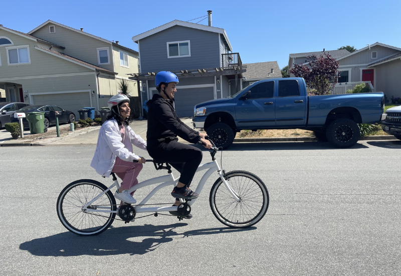 Two people riding a tandem bicycle together on a residential street.