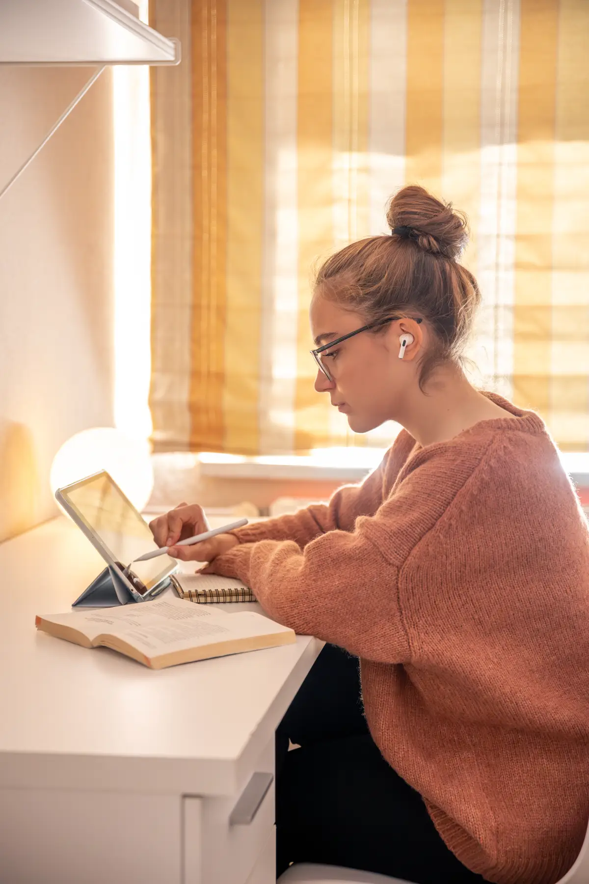 Mladá žena s brýlemi a drdolem studuje u stolu s tabletem, otevřenou knihou a poznámkovým blokem.
