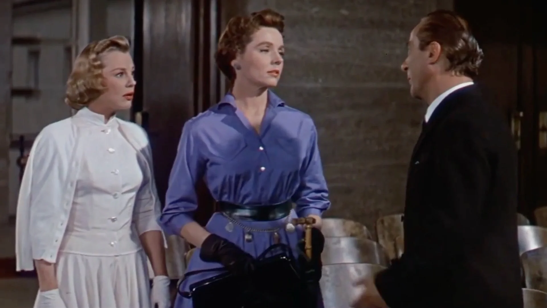 A man in a dark suit talks to two women dressed in vintage 1950s attire, one in a white dress and the other in a blue dress with black gloves.