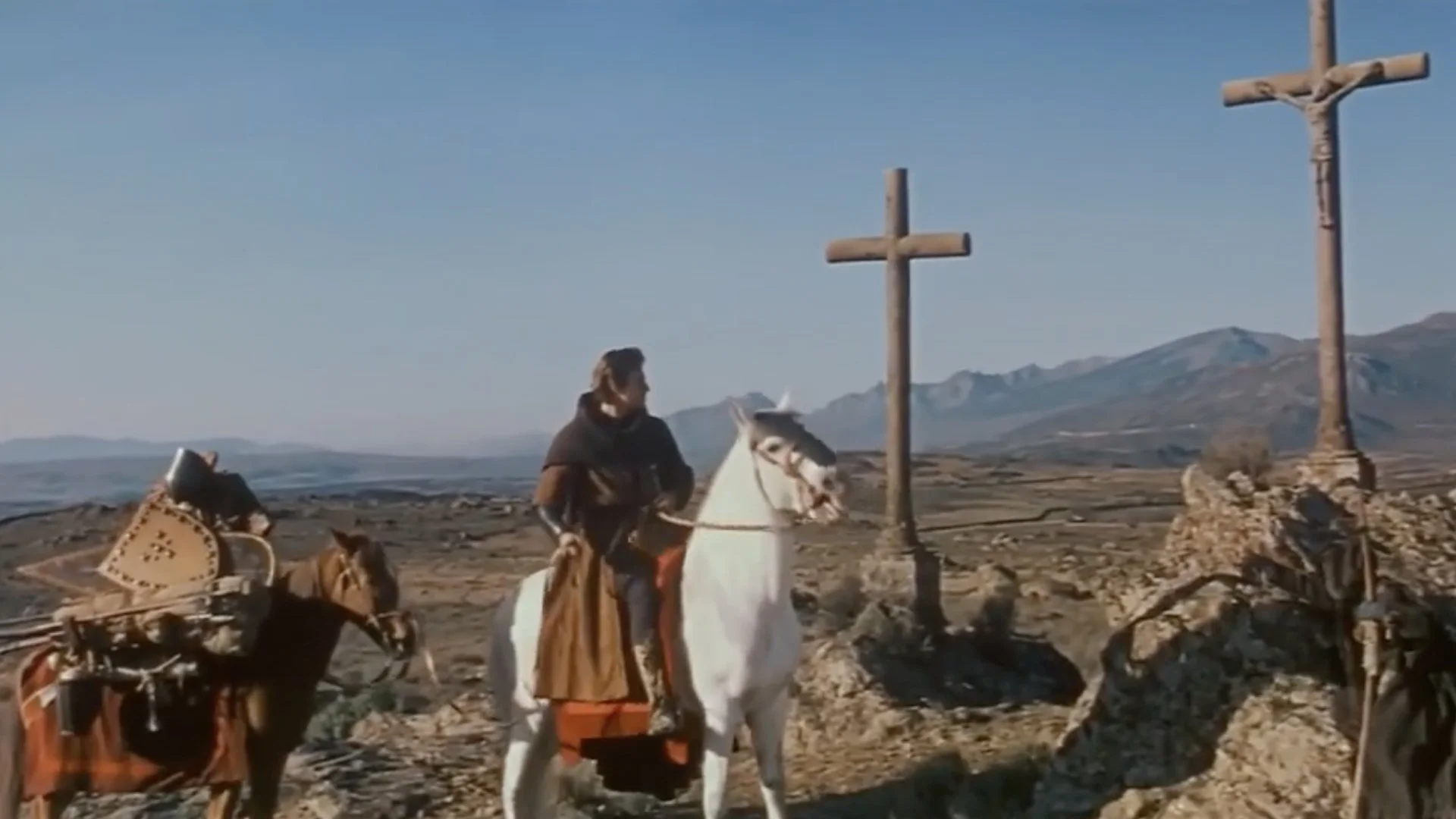 A medieval knight on a white horse looks to the right near two large stone crosses in a rocky, mountainous landscape.