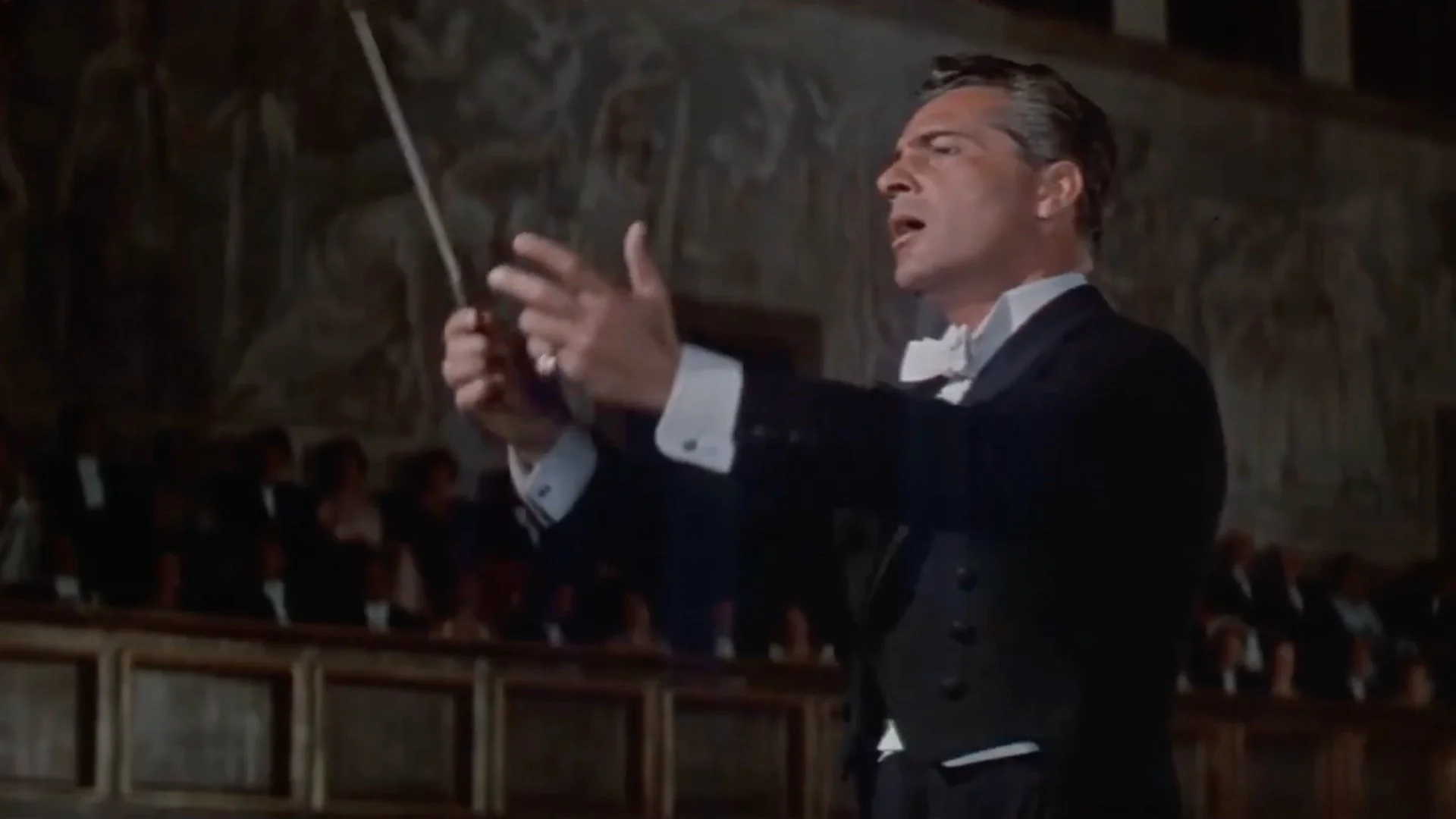 Man in formal attire passionately conducting an orchestra with a baton in a concert hall.