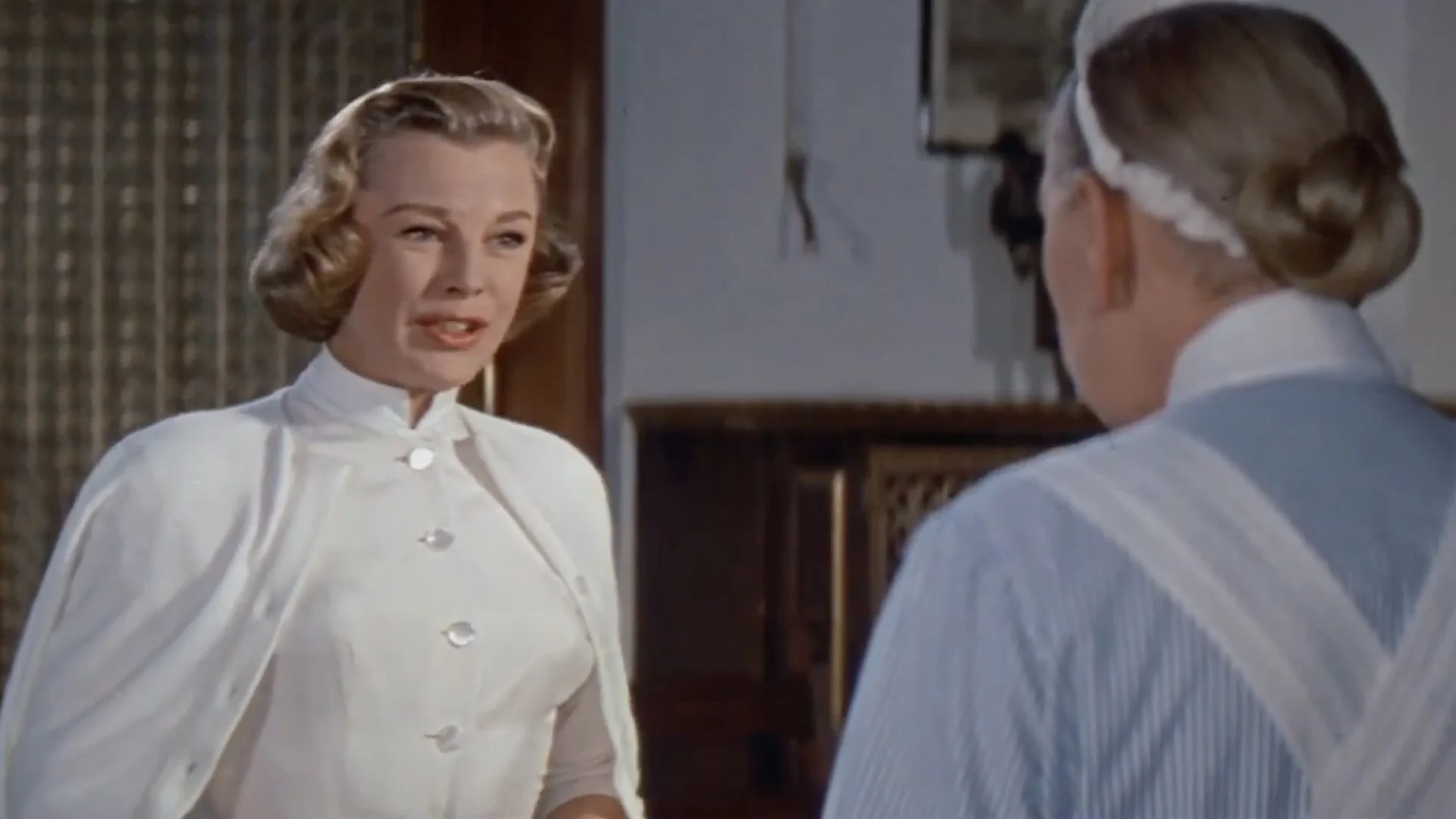Two women in vintage nurse uniforms facing each other in a room with wooden furniture.