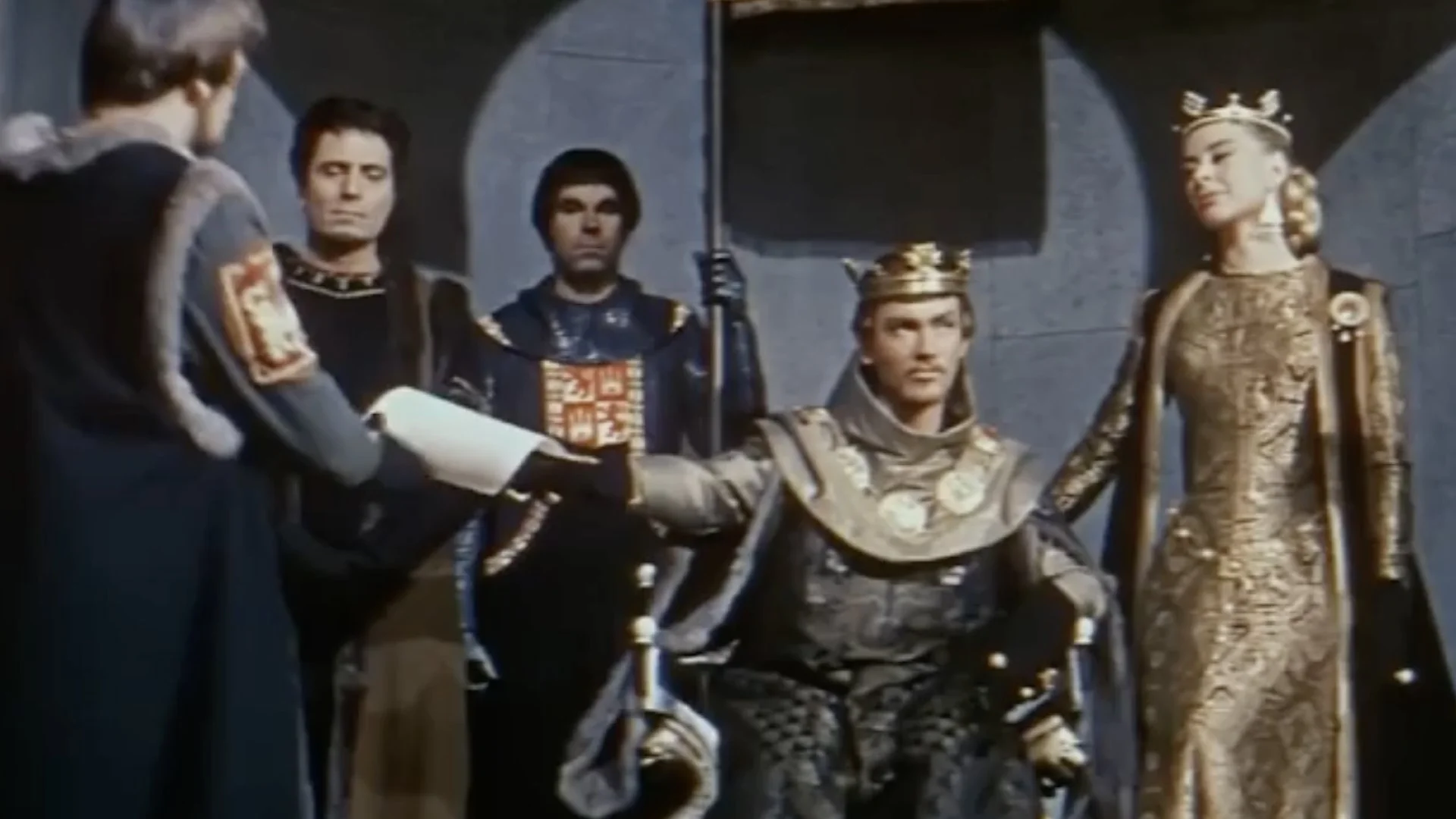 Medieval king in armor and crown seated on throne receiving a scroll from a standing man, with queen in gold dress and attendants behind.