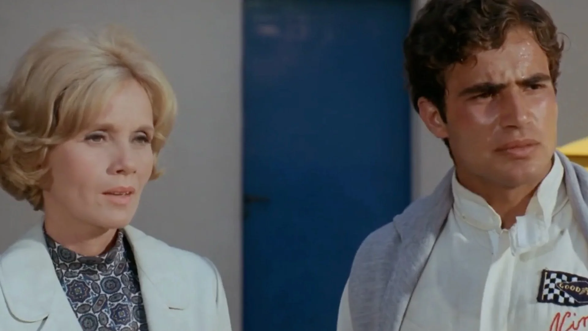 A woman with short blonde hair and a patterned blouse under a light-colored jacket stands next to a man in a white racing suit with a checkered patch and a gray sweater draped over his shoulders.
