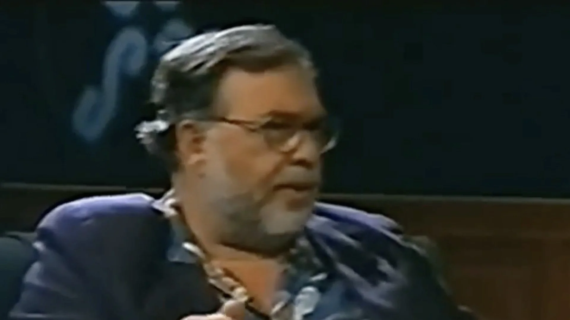 Older man with glasses and a beard speaking, seated in a dark setting.