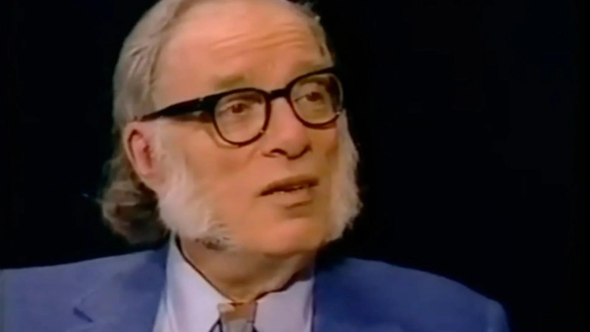 Elderly man with gray hair, sideburns, glasses, and a blue jacket looking to the side against a black background.