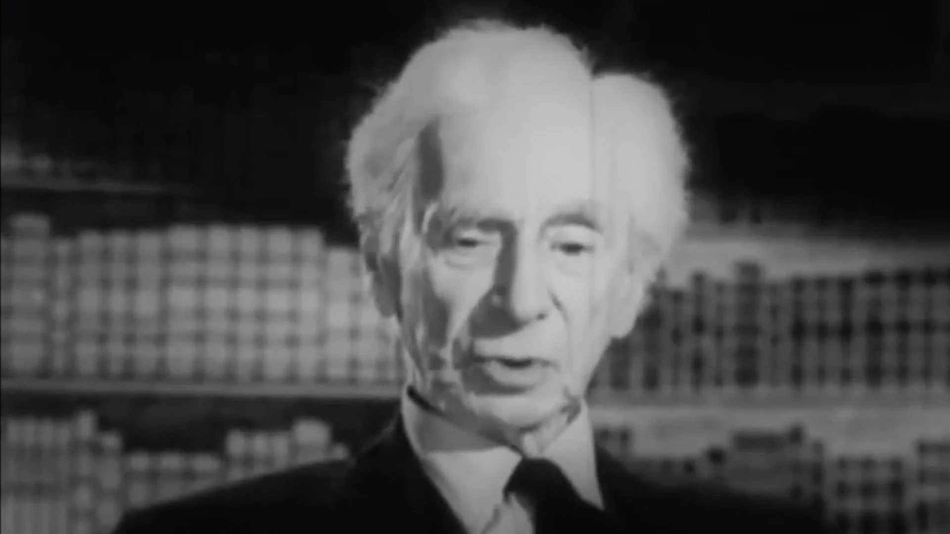 Black and white portrait of an elderly man with white hair speaking, with bookshelves in the background.