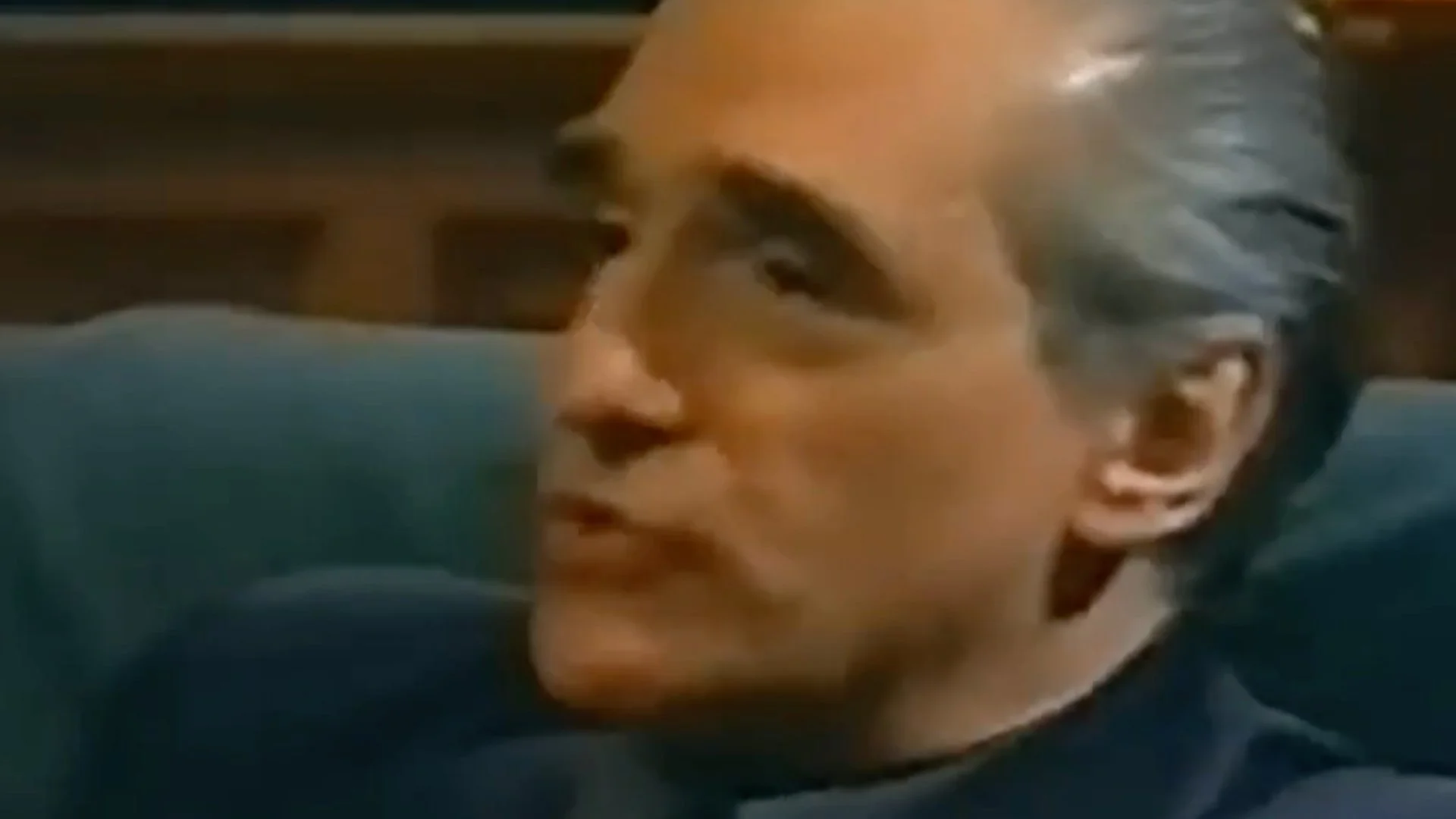 Close-up of a man with gray hair sitting and speaking, facing left.