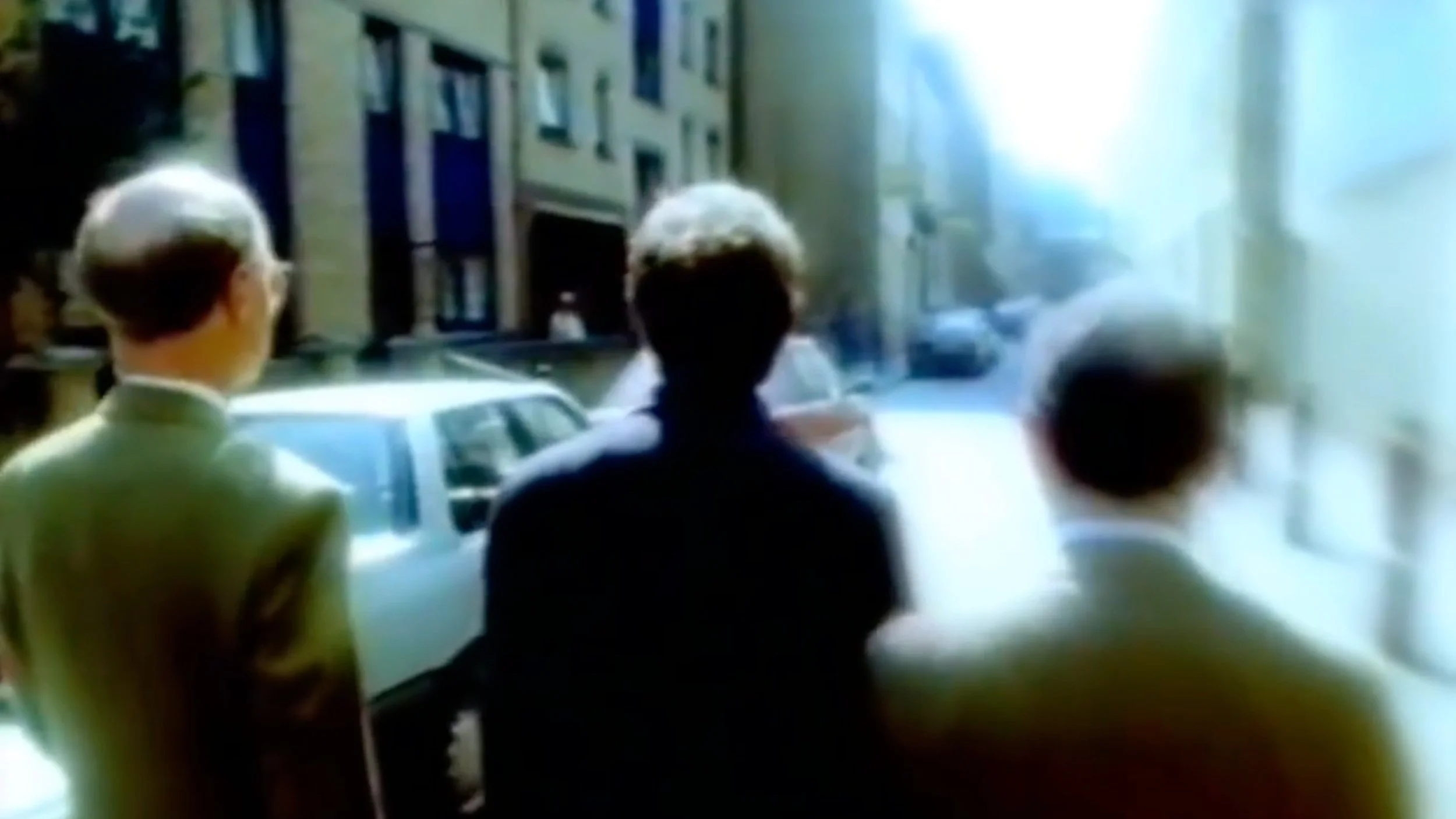 Three men standing on a street facing away, with cars parked along the side and buildings in the background.