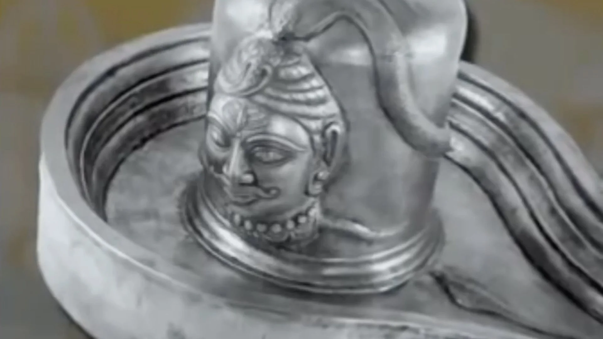 Close-up of a silver Shiva Lingam sculpture featuring a detailed face with traditional Indian ornamental headgear and jewelry.
