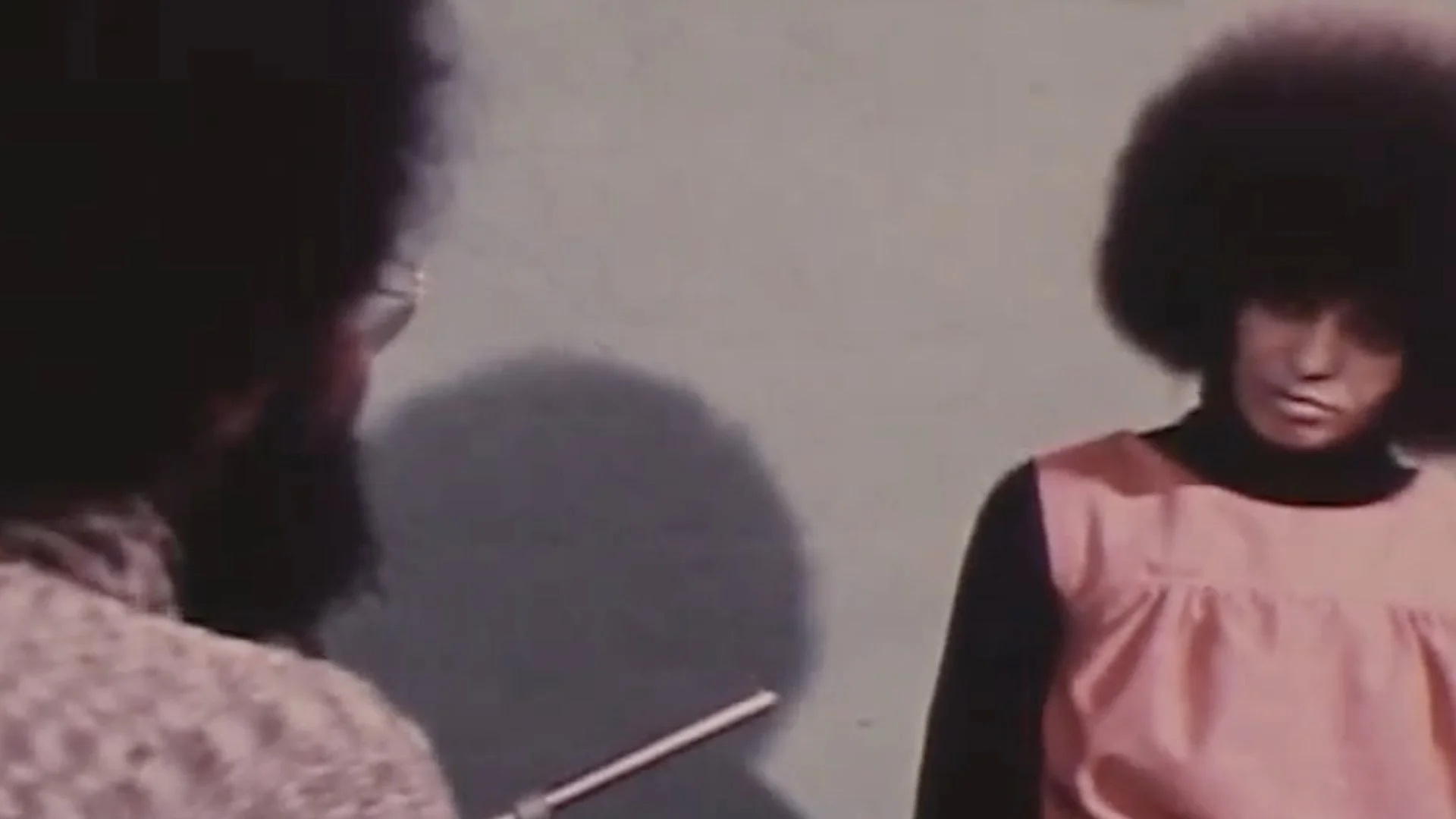 Two people in conversation; one with a beard facing away and one with an afro hairstyle wearing a pink dress looking down.