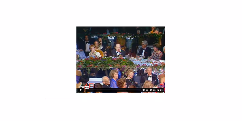People dressed formally sitting at round tables with festive floral centerpieces at a banquet or formal event.
