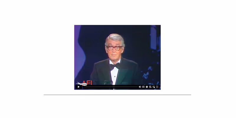 Man with gray hair and glasses wearing a tuxedo, speaking on stage against a dark purple background.