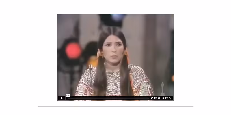 A woman with long dark hair wearing a patterned top and orange earrings is speaking in a video frame.