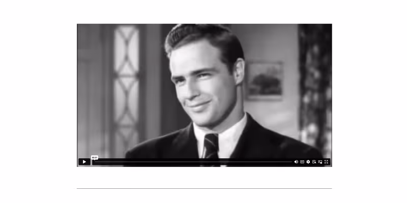 Black-and-white video still of a smiling man in a suit and tie.