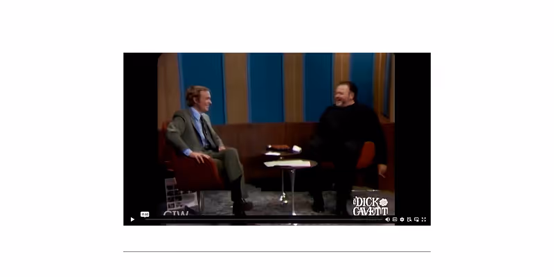 Two men sitting and laughing on a vintage talk show set with blue and brown panel background and a small round table between them.