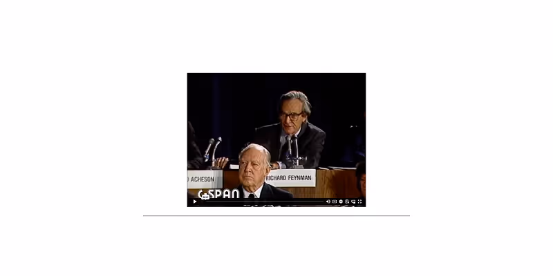 Richard Feynman speaking into a microphone at a panel event with another man seated in front.