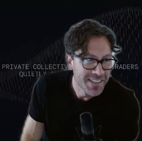 Man with glasses speaking into a microphone against a dark backdrop with faint text.