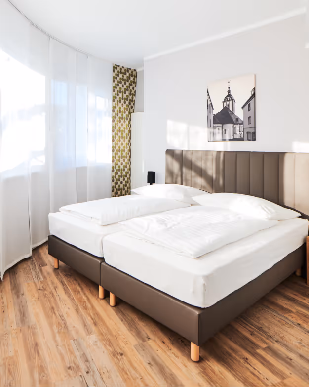 Bright bedroom with two single beds pushed together, white bedding, brown upholstered headboard, and wooden flooring.