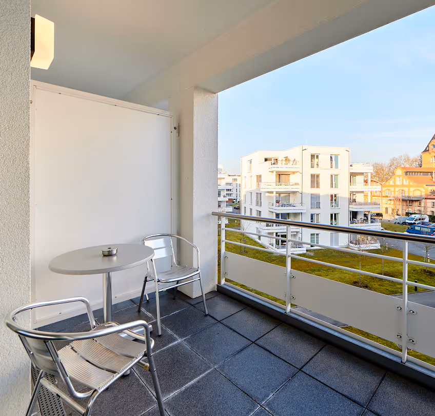 Balcony with two metal chairs and a small round table overlooking modern white apartment buildings and green lawn.