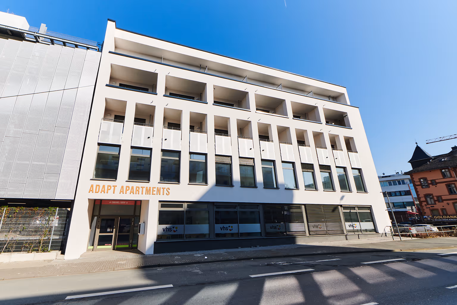 Modern white building with multiple windows and balconies labeled 'Adapt Apartments' under clear blue sky.