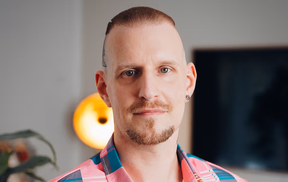Portrait of a man with a trimmed beard and short hair, wearing a colorful checkered shirt, in a cozy indoor setting.